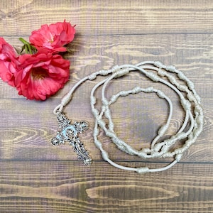 May include: White knotted rosary with a silver crucifix featuring a floral design and a figure of Jesus Christ. The rosary is on a wooden surface with pink flowers.