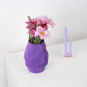 Puede incluir: Un jarrón morado texturizado contiene un ramo de flores rosas y rojas. Dos velas retorcidas de lavanda están en un pequeño soporte blanquecino. El jarrón tiene un diseño geométrico.