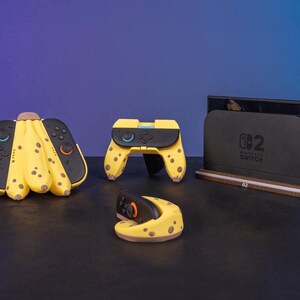 May include: A collection of Nintendo Switch accessories with a banana theme. Includes a Joy-Con controller grip, a Switch dock, and a Joy-Con charging dock, all in yellow with brown spots. The Switch dock has the text "Nintendo Switch" on it.