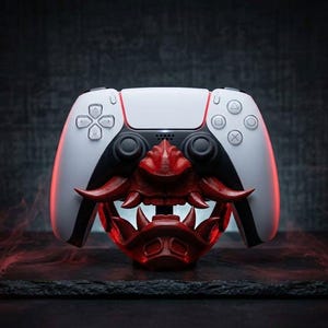 May include: A white and black PlayStation 5 controller with a red Oni mask design. The controller features a central touchpad, directional buttons, and action buttons. The red mask has sharp teeth and horns, adding a unique aesthetic to the gaming accessory.