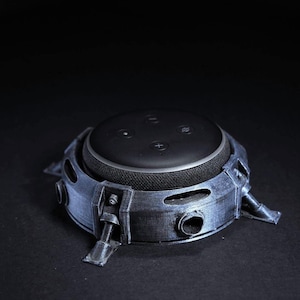 May include: A black smart speaker is housed in a metallic grey, industrial-style stand. The stand features a circular design with several protruding legs and circular openings. The speaker has control buttons on top.