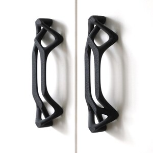 May include: Two black, abstract-shaped cabinet pulls mounted on a white surface. The pulls have a unique, open design with curved lines and geometric shapes. These decorative handles add a modern touch to any cabinet or drawer.