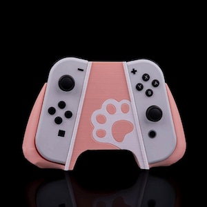 May include: A pink and white Nintendo Switch controller grip featuring a paw print design. The controller has black buttons and joysticks. The grip is designed to improve gaming comfort.