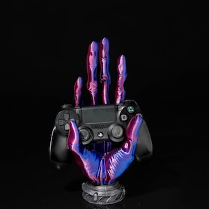 Könnte beinhalten: Ein schwarzer PlayStation 4-Controller wird von einem handförmigen Ständer gehalten. Der Ständer ist mit einem Farbverlauf aus lila und blauen Farbtönen bemalt. Der Sockel des Ständers ist dunkelgrau, und der Hintergrund ist schwarz.