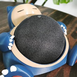 Puede incluir: Un soporte para altavoz con forma de Snorlax azul y beige con un altavoz de malla negra. El soporte tiene garras blancas y una barriga beige. El altavoz está encajado en el centro de la figura de Snorlax.