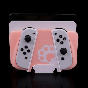 May include: A pink and white Nintendo Switch controller holder with a paw print design. The holder has a slot for the Switch console and is designed to hold the controllers. The controllers are gray and black.