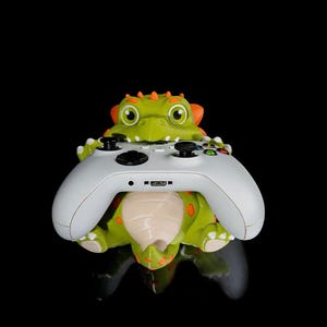 以下が含まれることがあります： 白のゲームコントローラーを抱える、緑とオレンジ色の恐竜型コントローラーホルダー。ホルダーにはオレンジ色のトゲと斑点があります。コントローラーには黒いボタンとUSBポートがあります。
