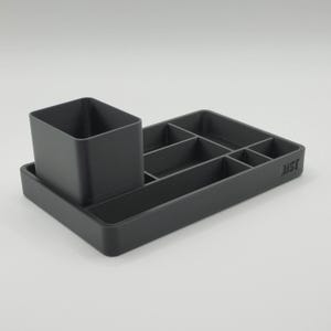 May include: A dark grey desk tidy with a pen pot and several compartments. The organiser has a rectangular base with divided sections for storing stationery. The pen holder is a square cup shape. The word "JUST" is printed on the side.