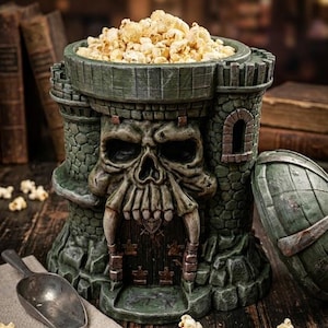 Könnte beinhalten: Eine grüne, burgförmige Popcornschüssel gefüllt mit Popcorn. Die Schüssel hat ein Totenkopf-Design und eine steinartige Textur. Ein Metalllöffel und eine Leinen-Serviette mit einem Totenkopf-Logo sind ebenfalls vorhanden.