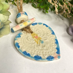 May include: A white ceramic heart-shaped pendant with blue and pink accents. The pendant has a detailed embossed design and is attached to a brown twine cord.