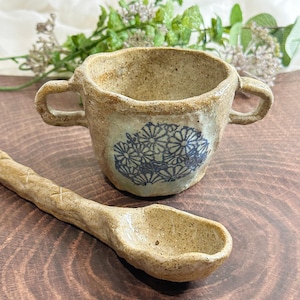 Può includere: Una piccola tazza in ceramica marrone rustica con due manici e un motivo floreale blu. La tazza è abbinata a un cucchiaio in ceramica marrone.