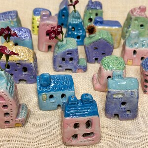 May include: A collection of miniature ceramic houses in various pastel colours, including pink, blue, green, and yellow. Each house has a unique design and features small windows and doors. Some houses have flowers or greenery on top.