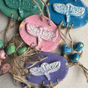 Puede incluir: Adornos de libélulas de cerámica hechos a mano en verde, azul, rosa y morado. Cada adorno ovalado presenta un diseño de libélula en relieve y está ensartado con hilo. También se incluyen cuentas decorativas en colores coordinados.