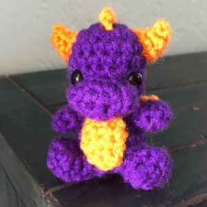 May include: A small crocheted purple dragon with orange horns and yellow crocheted accents. The dragon has black eyes and a cute expression.