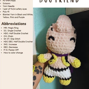 May include: A handmade crochet figure of a character from the Buu Friend series. The figure is primarily light pink with black, yellow, purple, and white accents. The text "BUU FRIEND" is at the top. The image also includes a list of supplies and abbreviations.