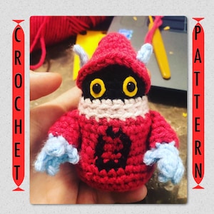 May include: Crocheted amigurumi toy in the shape of a red and black character with yellow eyes. The toy has a pink collar and blue arms.