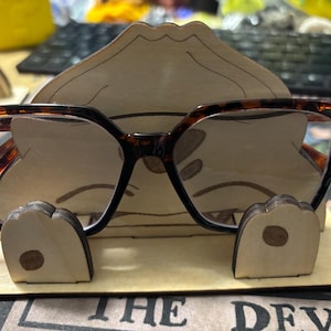 May include: A wooden eyeglasses stand shaped like a cartoon face with brown tortoise shell glasses resting on it. The stand has a light brown finish and the text "THE DEVIL" is visible at the bottom.