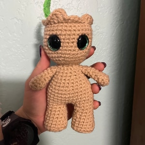 May include: A tan crocheted doll with large black eyes and a small green sprout on its head. The doll has a rounded body, arms, and legs. The doll is handmade and appears to be a character from a popular movie.