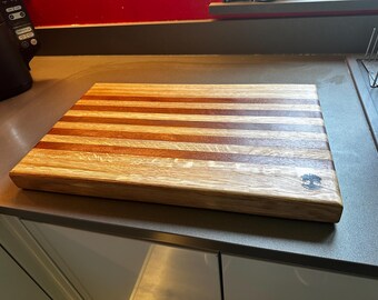 May include: A rectangular wooden cutting board with a striped pattern of light and dark wood. The board has a small, dark logo on the bottom right corner.