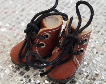 May include: A pair of miniature brown boots with black laces. The boots have a classic design with white stitching around the top and silver eyelets. The laces are tied in a knot at the top. The boots are likely for a doll or other small figure.