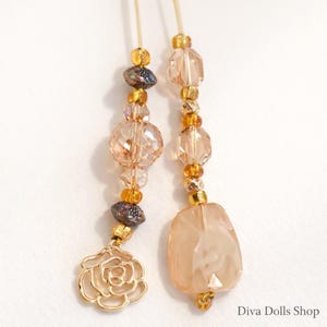 May include: Two beaded accessories with a rose charm and a rectangular pendant. The beads are in shades of gold, peach, and iridescent purple. The rose charm and other metal accents are gold. The text "Diva Dolls Shop" is in the bottom right corner.