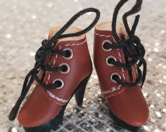 May include: A pair of miniature brown leather boots with black high heels and black laces. The boots have silver eyelets and white stitching around the top and sides. The heels are curved and the soles are black.