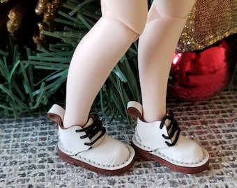 May include: A pair of miniature white ankle boots with black laces and brown soles worn on doll legs. The boots feature a pull tab at the heel, visible stitching along the base, and a rounded toe design. The footwear is constructed with a smooth white upper material and dark brown textured soles.