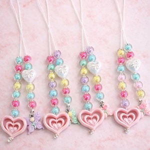 May include: Four beaded accessories with a pink heart pendant, butterfly charm, and a string of colorful beads. The accessories have a white heart-shaped bead and are strung on a white cord. The text "Diva Dolls Shop" is at the top.