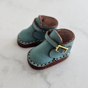 May include: A pair of miniature teal leather boots with brown soles and interior. Each boot features a strap with a gold-colored buckle and white stitching around the edges. The boots are designed for a doll or similar small figure.