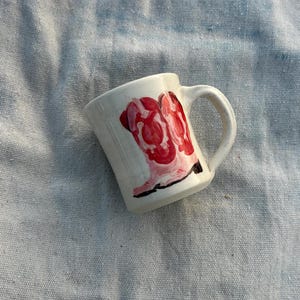 May include: A small, white ceramic mug with a curved handle. The mug features a hand-painted design of red and pink abstract shapes. The mug is set against a light blue, textured fabric background.
