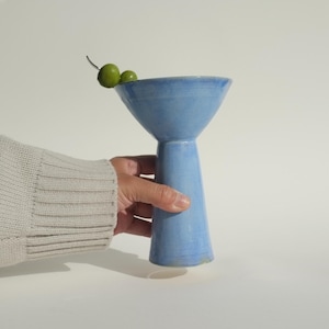 May include: A light blue ceramic cocktail glass with a wide, flared rim and a tapered base. Two green olives are skewered on a silver toothpick and rest on the rim of the glass. The vessel has a smooth, glossy finish and a solid, sturdy structure.