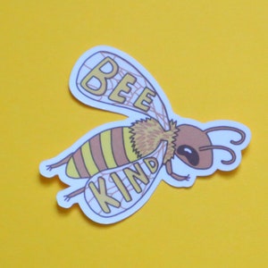 May include: A white sticker with a cartoon bee illustration. The bee has yellow and brown stripes and the words "Bee Kind" are written on the sticker.