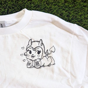 May include: White t-shirt with a black and white illustration of a cartoon devil with hearts around it.