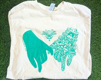 May include: A beige t-shirt with a green print of two hands holding a cluster of flowers. The text "Celebrate You" is printed on the shirt.
