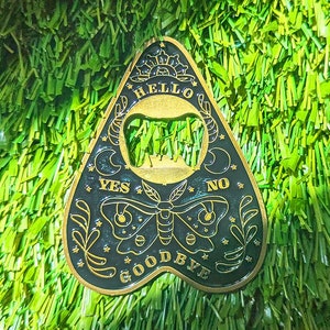 May include: A gold-colored metal ouija board charm with black lettering. The charm is shaped like a teardrop and features the words "Hello", "Yes", "No", and "Goodbye".