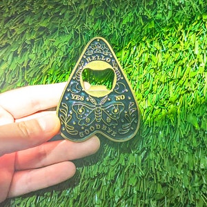 Puede incluir: Un abridor de botellas de metal negro y dorado en forma de tablero de ouija con las palabras "Hello", "Goodbye", "Yes", "No" y un dise&ntilde;o de luna creciente.