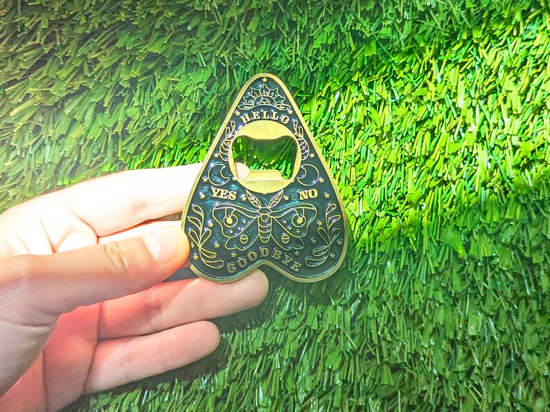 Puede incluir: Un abridor de botellas de metal negro y dorado en forma de tablero de ouija con las palabras "Hello", "Goodbye", "Yes", "No" y un dise&ntilde;o de luna creciente.