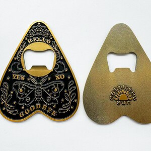 Puede incluir: Un abridor de botellas de metal dorado en forma de coraz&oacute;n con un dise&ntilde;o negro y dorado. El dise&ntilde;o presenta una mariposa, estrellas y las palabras "Hello", "Yes", "No" y "Goodbye".
