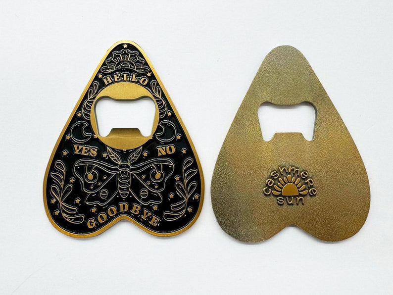 Puede incluir: Un abridor de botellas de metal dorado en forma de coraz&oacute;n con un dise&ntilde;o negro y dorado. El dise&ntilde;o presenta una mariposa, estrellas y las palabras "Hello", "Yes", "No" y "Goodbye".