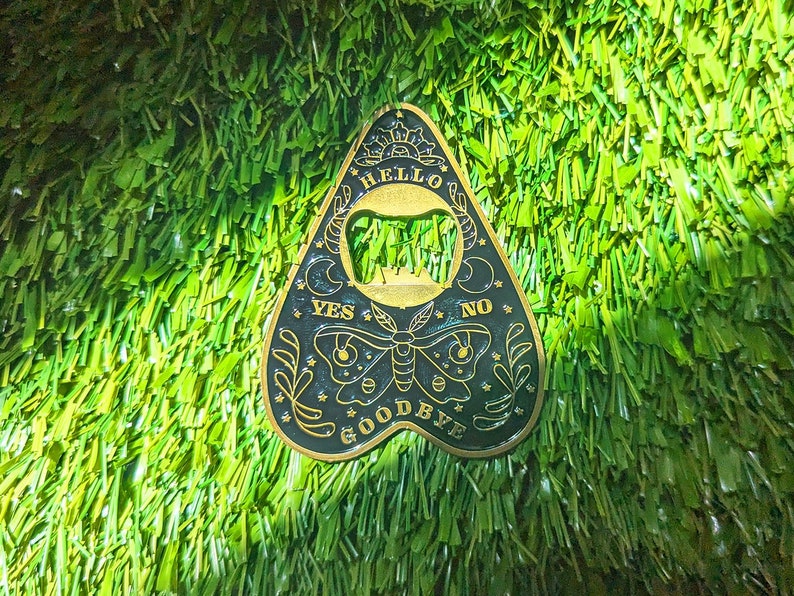 Puede incluir: Un amuleto de tablero de ouija de metal dorado con letras negras. El amuleto tiene forma de l&aacute;grima y presenta las palabras "Hello", "Yes", "No" y "Goodbye".