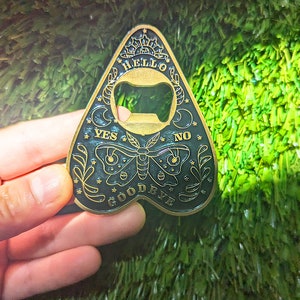 Puede incluir: Un abridor de botellas de color dorado con forma de planchette de Ouija. La planchette tiene un fondo negro con una mariposa blanca, estrellas y las palabras "YES", "NO", "HELLO", "GOODBYE".