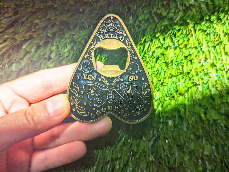 Puede incluir: Un abridor de botellas de color dorado con forma de planchette de Ouija. La planchette tiene un fondo negro con una mariposa blanca, estrellas y las palabras "YES", "NO", "HELLO", "GOODBYE".