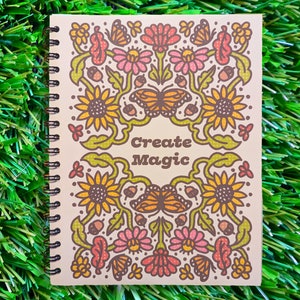 May include: A cream-colored spiral bound notebook with a floral design featuring sunflowers, butterflies, and acorns. The text "Create Magic" is printed in brown on the front cover.
