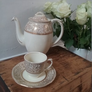 May include: A white teapot and matching teacup and saucer with a gold floral pattern. The teapot has a lid and a spout. The teacup and saucer are on a wooden surface.
