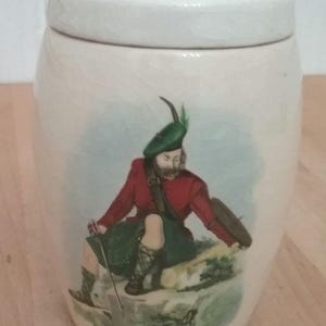May include: White ceramic jar with a lid featuring a painting of a person wearing a red jacket, green kilt, and a green hat with a feather. The person is holding a stick and standing on a rock.