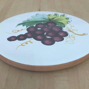 May include: Circular white ceramic plate featuring a hand-painted grape design. The grapes are dark purple, with green leaves and yellow vines. The plate has a brown rim.