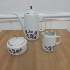 May include: A white porcelain tea set with gold trim, including a teapot, creamer jug, and sugar bowl. Each item is adorned with a floral design of pink, blue, and purple roses. The teapot is about 23 cm tall.