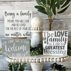 May include: A tiered tray with a white background and a farmhouse style. The tray is decorated with small wooden signs with inspirational quotes about family. The signs are in gray and black lettering. The tray is decorated with greenery.
