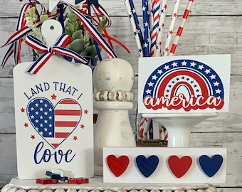 May include: A tiered display of patriotic decorations. Includes wooden signs with phrases like "Land That I Love," "America," and "Stars and Stripes." Red, white, and blue stars, hearts, and decorative straws complete the festive arrangement.
