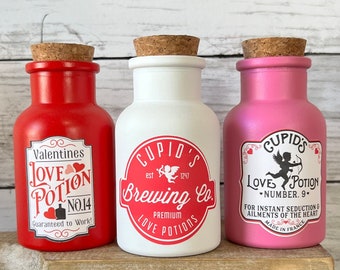 May include: Three glass bottles with cork tops and labels. The bottles are red, white, and pink. The labels have a Valentine's Day theme and say "Love Potion" with different numbers. The bottles are sitting on a wooden shelf.